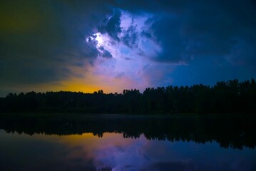 lightning strikes at night behind the clouds in the reflection of the lake in autumn