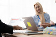 © Kritdanai - Collaborative Discussion: A diverse team collaborates on a project, with a young woman in a hijab confidently presenting her ideas in a modern office setting.