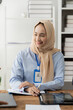 © Kritdanai - Confident and Focused: A young businesswoman wearing a hijab sits at her desk, her smile radiating confidence and focus as she works on a project. She exudes professionalism and success, representing