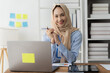 © Kritdanai - Confident and Ready:  A young businesswoman, wearing a hijab, smiles confidently while holding a pen and looking directly at the camera.  She is seated at her desk with a laptop, a tablet, and a stack