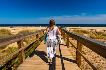 Wall Mural - Long-haired woman wearing white top and white pants with colorful patterns walking to sandy beach and carrying bodyboard on sunny day on wooden promenade. Vacation in Algarve, Portugal. Back view