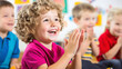 © Nataliia - Happy young boy with curly hair clapping hands in a classroom, surrounded by other cheerful children
