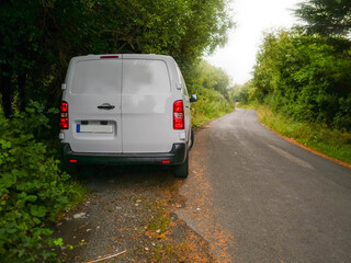 Wall Mural - A white medium size van is parked on a small county road in rural area with a red tail light. The road is empty and the sun is shining brightly. Product delivery to remote locations concept.