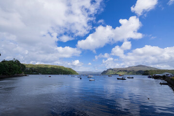 A serene landscape featuring a body of water with boats and buildings along the shore. The image captures a peaceful scene with a cloudy sky overhead.  Portree, Scotland, isle of skye