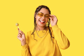 Wall Mural - Beautiful young African-American woman with dreadlocks holding lollipop and showing tongue on yellow background