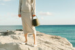 © Ulas&Merve/Stocksy - Woman Holding Bag on Rocky Seashore with Blue Ocean Background
