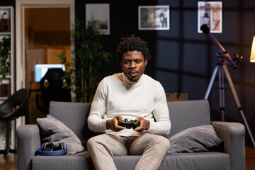 Wall Mural - Player using controller to play videogame on gaming console in cozy apartment. African american gamer using high tech gamepad to destroy enemies, relaxing at home on couch