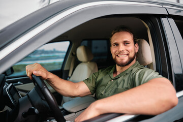 Wall Mural - Smiling man in a green shirt sitting in a car, posing through the open driver s window, with a blurred urban background