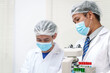 © NanSan - Two Asian scientists in lab coats sit at a table in a laboratory, examining samples through a microscope. The room is equipped with scientific tools, liquids, and chemistry materials.