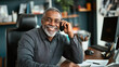 © altitudevisual - Smiling middle-aged man with gray hair and beard talking on a phone sitting at his office desk with a computer, notepad, and documents.