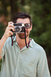 © JoseIMartin - A man smiles while taking a photo with a vintage camera in a sunny park. He wears sunglasses and enjoys the outdoor setting, capturing the moment.