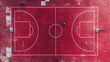 © Thamrong - a drone captures an aerial view of a deep red basketball court with white lines. The court is empty except for a few players practicing at one end. The red surface of the court stands out boldly