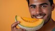 © Irina Kozel - A young Latino man smiles while holding a slice of cantaloupe close to his mouth against a light orange background