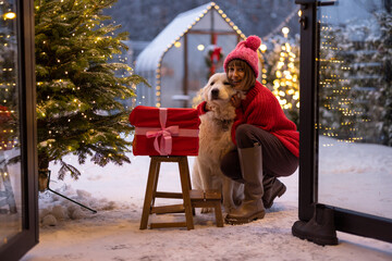 Woman hugs with her adorable dog near Christmas tree at beautifully decorated snowy yard, celebrating and having fun together on winter holidays. Concept of happy winter holidays, magic and love