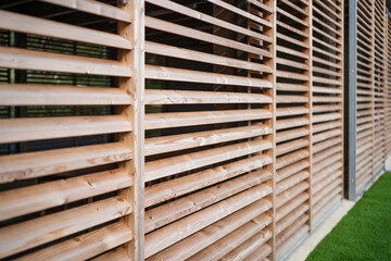 Wooden slats with a close-up view form a patterned, textured outdoor structure, with natural light and greenery visible through the gaps, showcasing the design and alignment of each wooden plank.