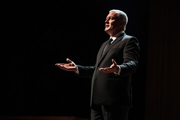 A man dressed in a formal black suit stands on stage, arms extended as he delivers a passionate speech under dramatic lighting