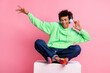 © deagreez - Full body portrait of cool young man sit cube headphones dance wear green hoodie isolated on pink color background
