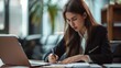 © COK House - A focused young woman in office attire sits at a desk, writing with a pen and using a laptop. Books are nearby, showcasing her diligent work ethic.
