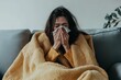 © Денис Никифоров - Woman sitting on a couch wrapped in a yellow blanket, sneezing into a tissue, suffering from cold or flu symptoms and seeking comfort.