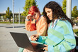 © Seventyfour - Waist up shot of multiethnic teenage girl with black curly hair pointing at laptop screen, while using computer together with female friend on bench in sunlit city park