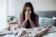 © Денис Никифоров - Woman sitting in bed sneezing into a tissue, covered in a cozy blanket, feeling unwell from flu or cold symptoms.
