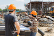 © skarie - Two construction workers discuss project details on-site, using a tablet and blueprints to plan the next steps amid the bustling construction environment.
