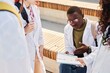 © Seventyfour - Medium shot of young male African American med student in white coat reviewing material in notebook discussing notes with classmates while studying outdoors on campus