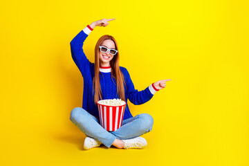 Wall Mural - Photo of young woman in blue stylish pullover and jeans sitting alone eating popcorn pointing fingers mockup isolated on yellow color background