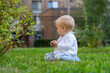 © sommersby - A young child sits on the grass, curiously examining plants nearby in a green park during daylight surrounded by urban scenery