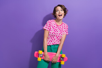 Poster - Photo of adorable cute girl wear stylish pink clothes hold skateboard look up empty space isolated on purple color background