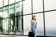 © BGStock72 - Businesswoman in a light gray suit smiles while walking past a modern glass building on a sunny day, engaged in a lively phone conversation