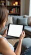 © umut hasanoglu - Woman holding a tablet while sitting in a cozy living room with a blank screen.