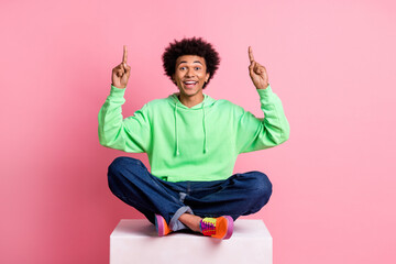 Full size photo of nice young man direct fingers up empty space sit podium wear green hoodie isolated on pink color background