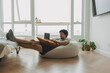 © Sevendeman - Asian man working with his laptop lying on the bean bag sofa in living room.