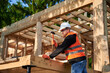 © anatoliy_gleb - Carpenter constructing wooden frame house near forest. Bearded man with glasses hammering nails into structure, wearing protective helmet, construction vest. Concept of modern ecological construction.
