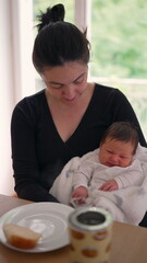 Mother holding her baby wrapped in a white blanket, sitting at a dining table with a piece of bread and a coffee mug, sharing a peaceful moment