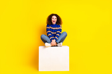 Wall Mural - Full size photo of overjoyed woman wear knit sweater jeans sit on white cube legs crossed laugh isolated on yellow color background