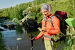 © pressmaster - Mature female hiker with smartphone, trekking sticks and backpack looking through mobile app with map or navigator