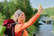 © pressmaster - Side view of mature female hiker in activewear looking at smartphone screen while taking selfie in natural environment