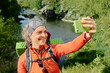 © pressmaster - Cheerful mature woman in activewear and headband looking at screen of smartphone while taking selfie or recording video on trip