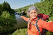 © pressmaster - Active mature female blogger and hiker pointing at riverside while looking at smartphone camera during livestream on summer trip