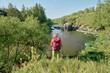 © pressmaster - Rear view of mature female hiker with backpack standing in front of wide and long river flowing between banks with green trees and grass