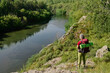 © pressmaster - Long shot of mature female hiker or camper with backpack and trekking sticks admiring riverside and natural landscape during trip