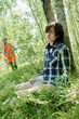 © pressmaster - Young unconscious female camper in casualwear sitting by tree on the ground covered with green grass against lifeguard in safety vest
