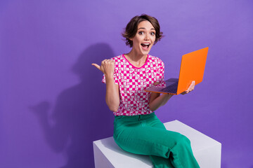 Wall Mural - Photo of charming impressed woman sit on platform with laptop in arm indicating at sale empty space isolated on violet color background