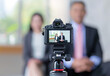 © MD Media - Close-up of a camera on a tripod, with two business people sitting in the background, A camera is placed on the tripod, with two business people standing in front of it for an interview video