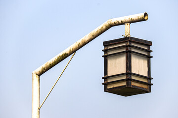 Close-up of a lantern-shaped street light, mounted on a rusted metal pole, set against a clear blue sky. The aged design and rust highlight its vintage aesthetic