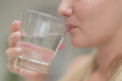 © ultramansk - A beautiful European woman smiles happily, holding a glass of water, drinking natural mineral water from a clear glass, inside a residential. A healthy woman shows her face, complete physical health.