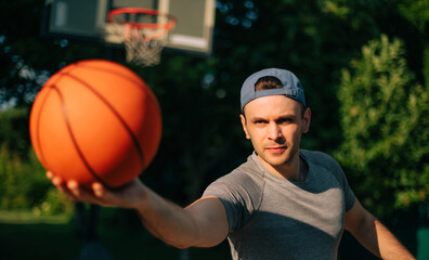 Wall Mural - Focused man in a cap extends a basketball toward the camera, creating a dynamic, action shot. The outdoor background, with a hoop in soft focus, highlights his intense, determined expression