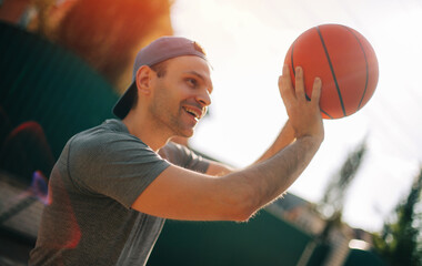 Wall Mural - A smiling man in a backward cap prepares to shoot a basketball, with warm sunlight highlighting his face. The vibrant orange ball contrasts with the soft focus of the outdoor background